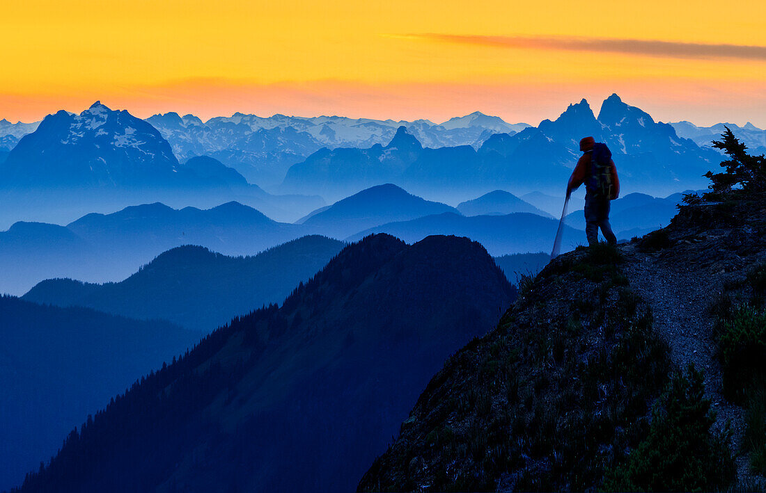USA, Staat Washington. Ein Backpacker, der bei Sonnenuntergang von der Skyline Divide in den North Cascades in der Nähe von Mt. Baker absteigt. Prominente Gipfel sind Judge Howay und Robie Reid in Kanada. Digitaler Verbund. (HERR)