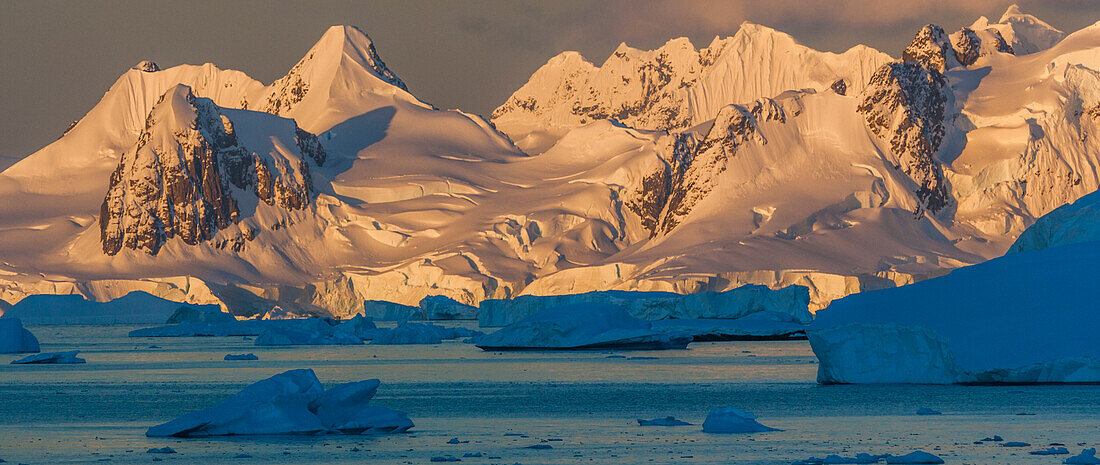 Morning light shines on the mountains of Antarctica, while the icebergs in the ocean remain shadowed.