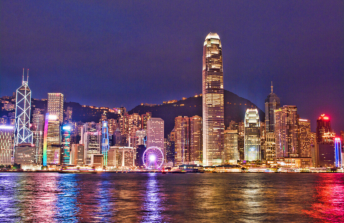 Hong Kong China skyline harbour night with new Ferris Wheel in city skyscrapers and reflections on water and mountain peak in background