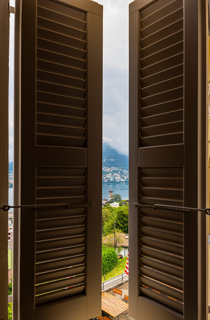 Window View with Shutter with Tower in Cityscape and Mountain and Lake Lugano in a Cloudy Day in Ticino, Switzerland.