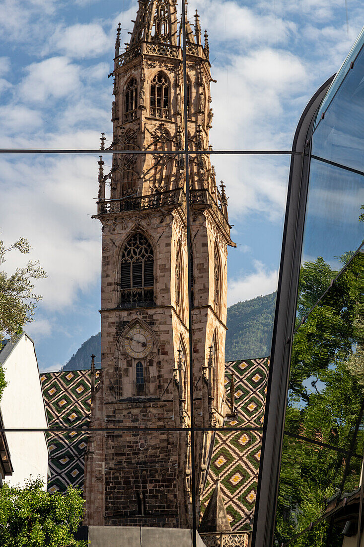 Bell tower of the Cathedral reflected in a glass building, under clouded sky Bolzano, South Tyrol, Italy