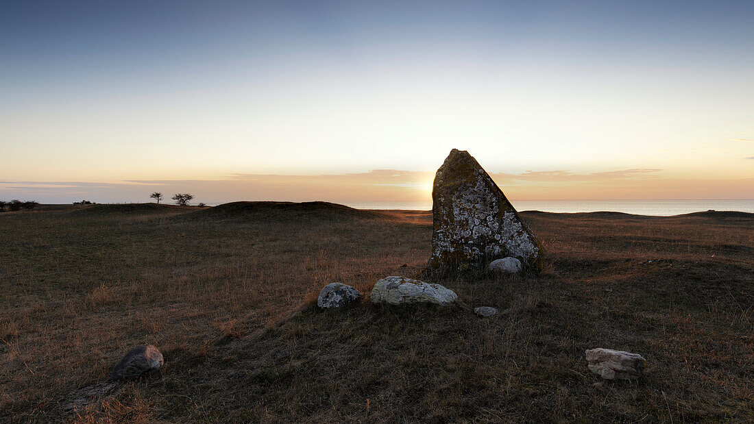 Tombstone and smaller stones on hilly meadow. Sun sets behind old tombstone. Ottenby burial ground, Degerhamn, Öland, Sweden.