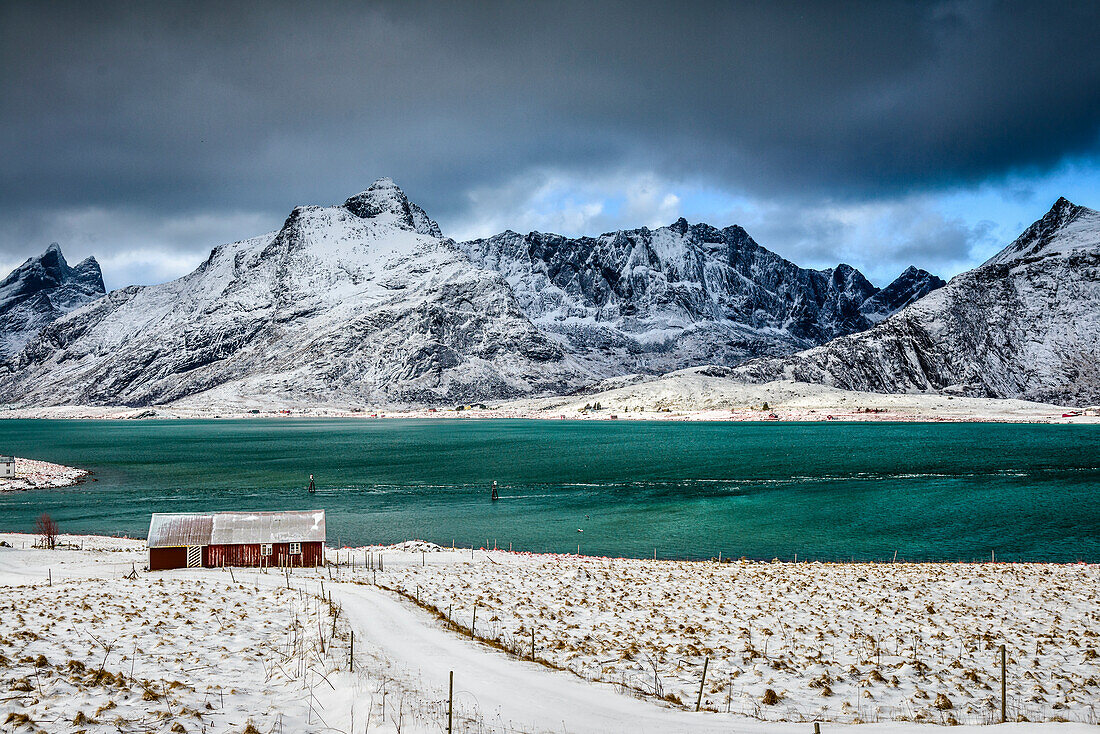 Die Lofoten-Inseln im Schnee, türkisgrünes Wasser, kleines Fischerdorf und Häuser am Ufer unter steigenden Bergen.