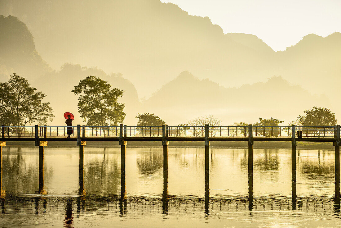 One person on a footbridge above water, mountains in the mist at dawn.