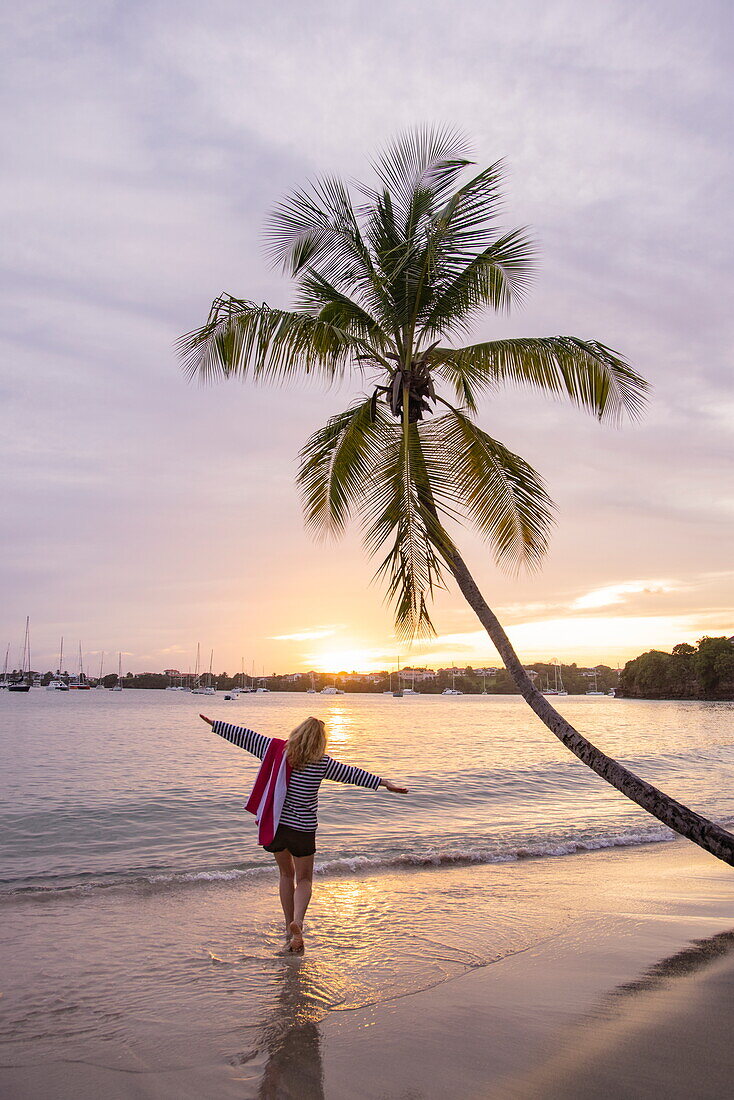 Frau streckt ihre Arme aus, Strand von L'Anse Aux Epines mit schiefer Kokospalme bei Sonnenuntergang, in der Nähe von Saint George's, Saint George, Grenada, Karibik