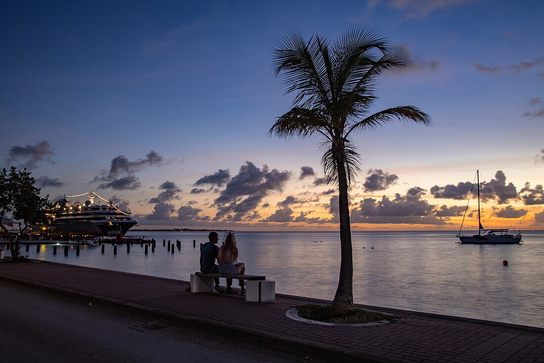 Paar auf Bank, Palme, Segelboot und Expeditionskreuzfahrtschiff World Voyager (nicko cruises) am Pier bei Sonnenuntergang, Kralendijk, Bonaire, Niederländische Antillen, Karibik