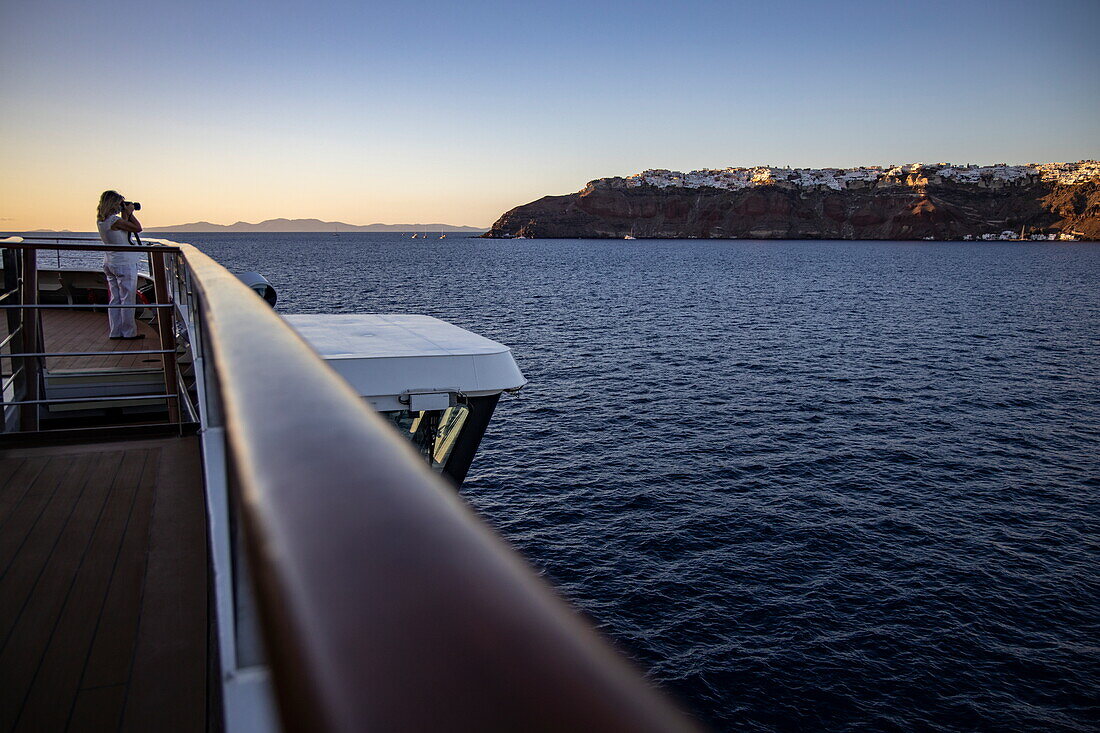 Railings of expedition cruise ship World Explorer (Nicko Cruises) and woman photographing the houses on the cliffs in the distance, Santorini, South Aegean, Greece, Europe