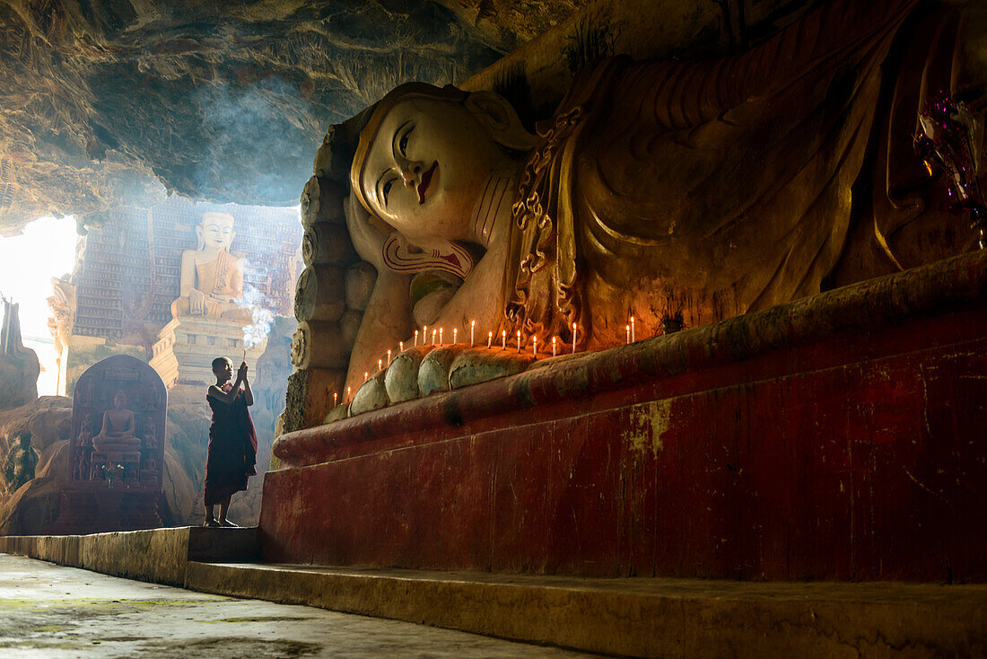 Ein Höhlentempel, eine große liegende Buddha-Statue, brennende Kerzen und ein betender Mönch, Myanmar, Asien