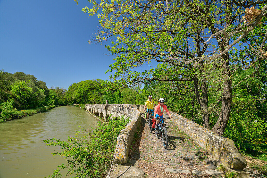Two people ride bikes across bridge on Canal du Midi, Pont-Canal de l'Argent Double, Canal du Midi, UNESCO World Heritage Canal du Midi, Occitania, France