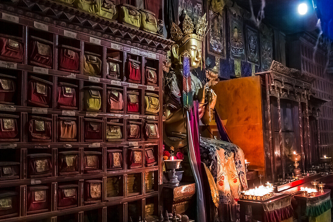 Buddha-Figur und antike Schrankwand mit Kerzen und Opfergaben im dunklen Inneren einer Gebetshalle im Kloster Kumbum Champa Ling, Qinghai, China