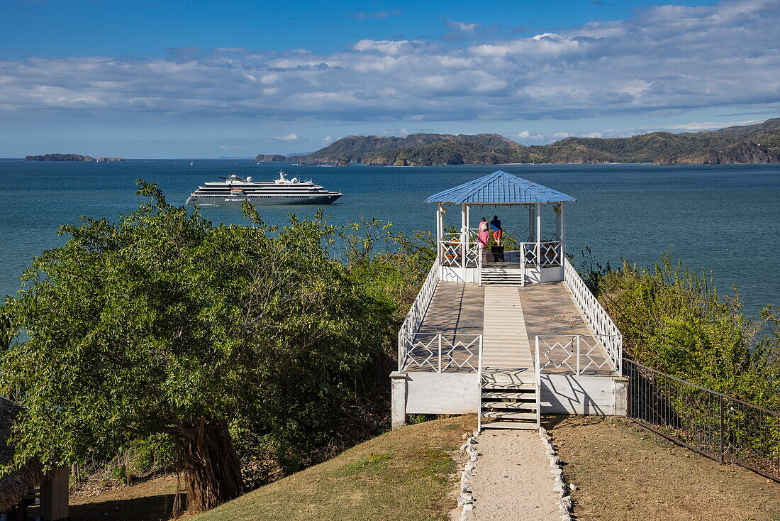 Paar im Pavillon des Dunama Casino & Hotel mit Expeditionskreuzfahrtschiff World Voyager (nicko cruises) in der Bucht dahinter, Playa Flamingo, Guanacaste, Costa Rica, Mittelamerika