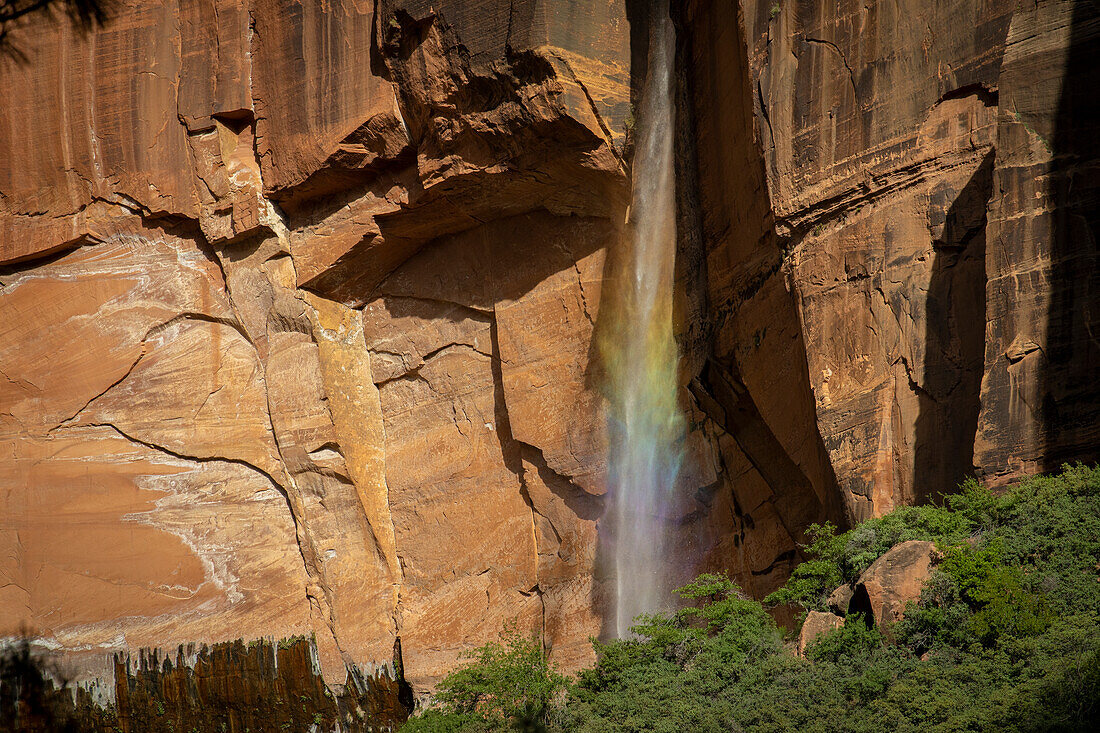 Small waterfall in front of a red rock face is illuminated by the sun and shines in rainbow colors.