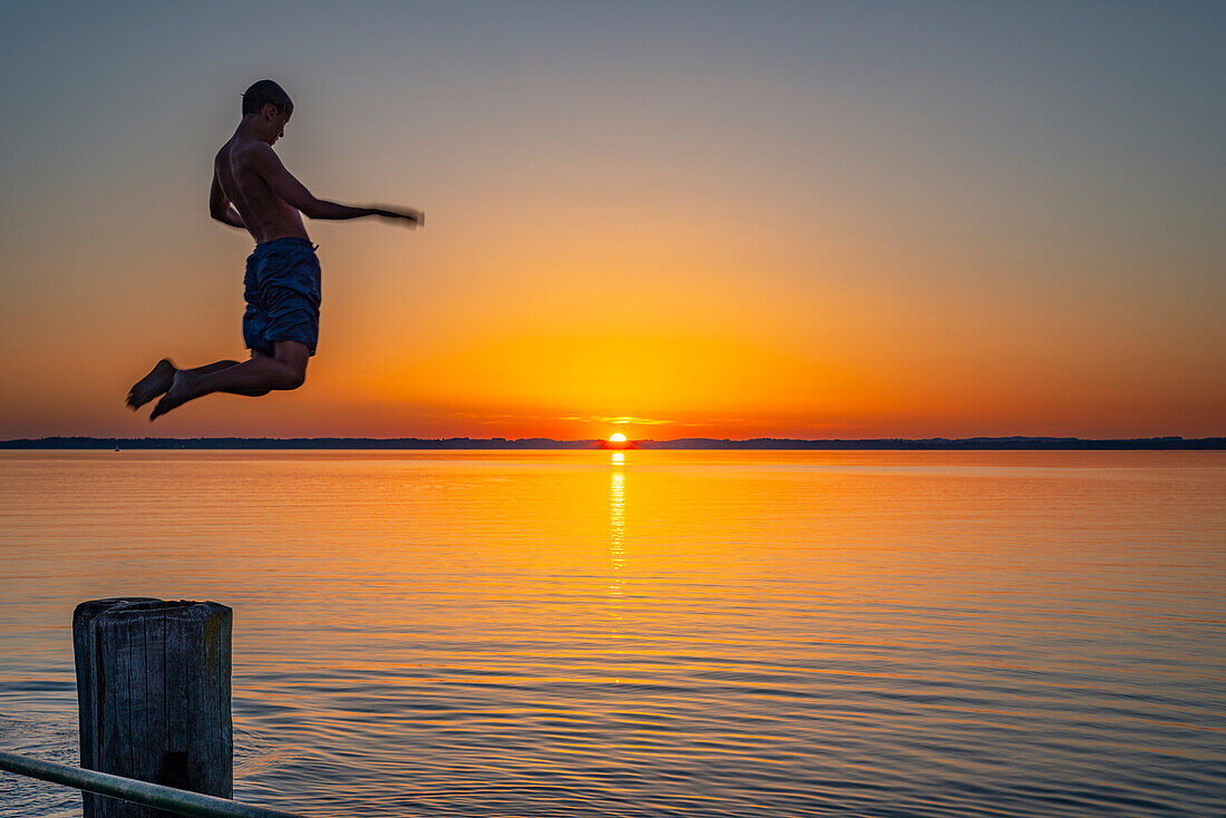 Boy jumping from the wooden pier into the water at sunset, Chieming, Chiemsee, Chiemgau, Bavaria, Germany, Europe