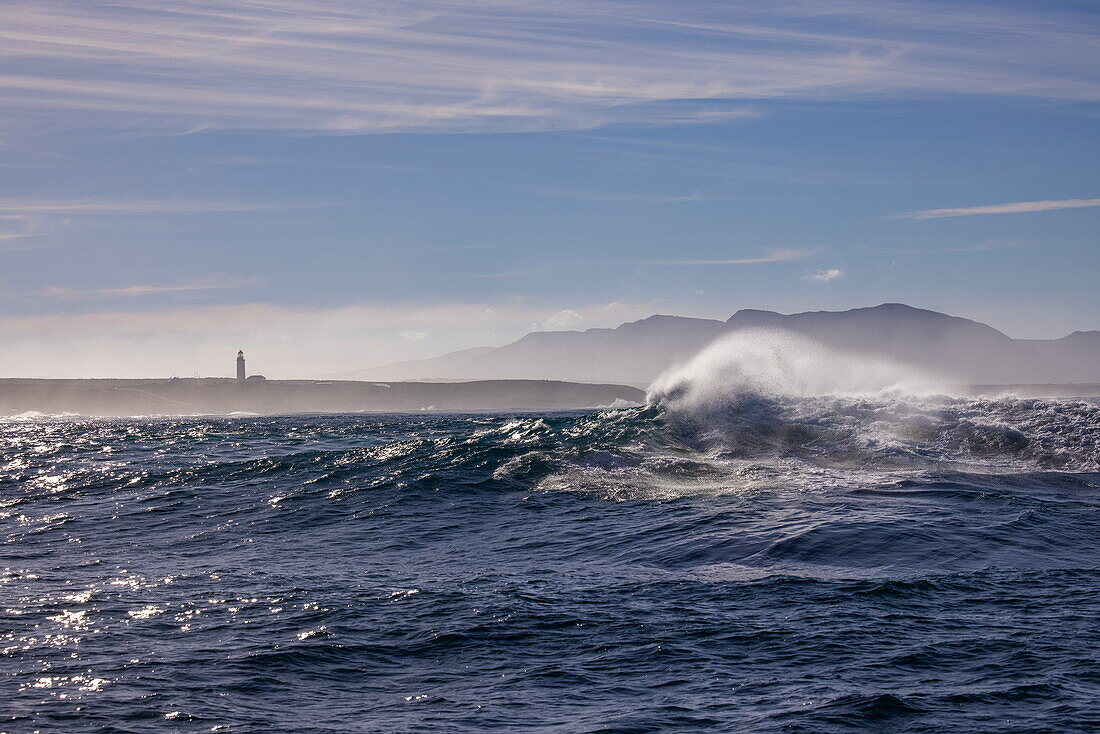Brechende Welle im Südatlantik mit Blick zum Leuchtturm Danger Point Lighthouse, in der Nähe von Gansbaai, Westkap, Südafrika