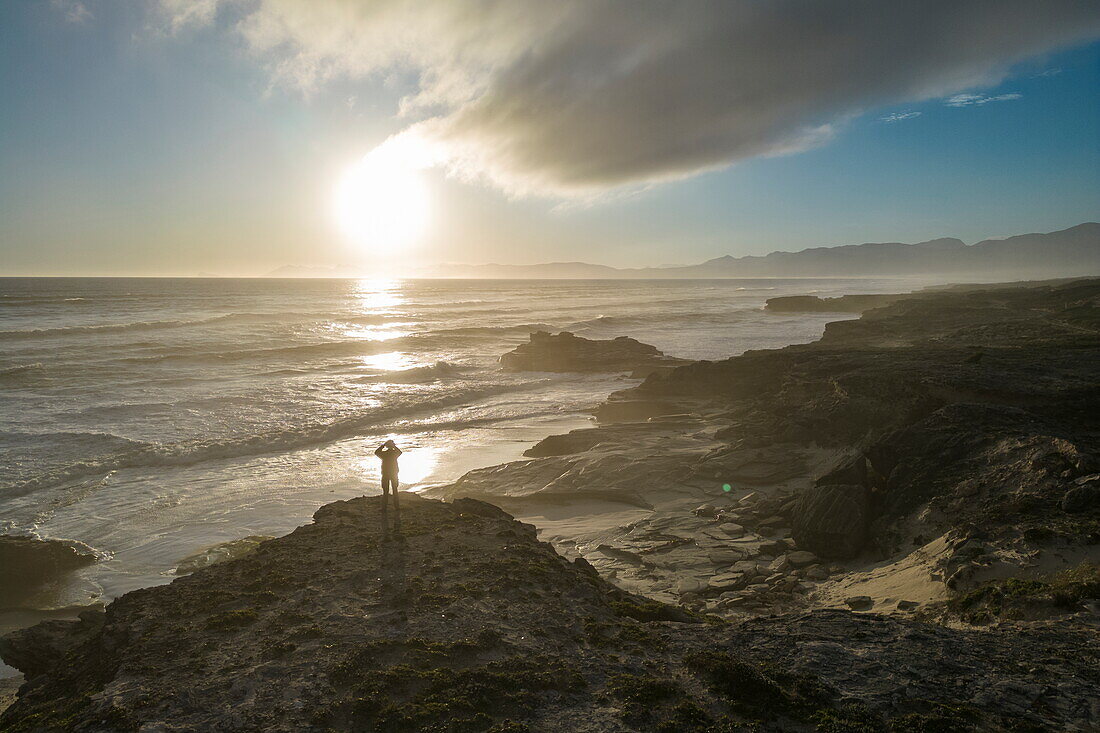 Luftaufnahme von einem Mann auf einem Felsvorsprung bei Sonnenuntergang am Strand, im Walker Bay Nature Reserve, Gansbaai De Kelders, Westkap, Südafrika