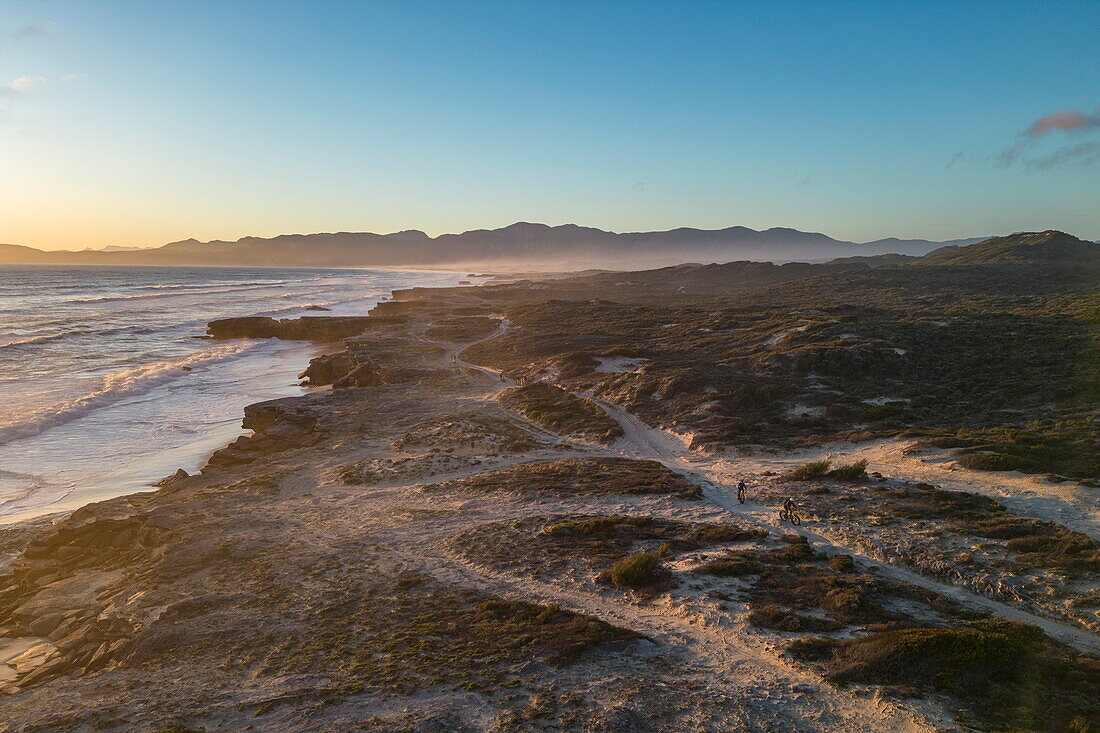 Luftaufnahme von Radfahrern mit Fat Tire Fahrrädern bei Sonnenuntergang auf Sandwegen entlang der Küste und am Strand, im Walker Bay Nature Reserve, Gansbaai De Kelders, Westkap, Südafrika