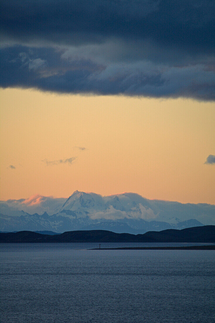Argentina; Province of Tierro del Fuego; on the border with Chile; Beagle Channel; Snow capped mountain peaks of Isla Navarino in Southern Chile; dark rain clouds in the orange sky of the setting sun