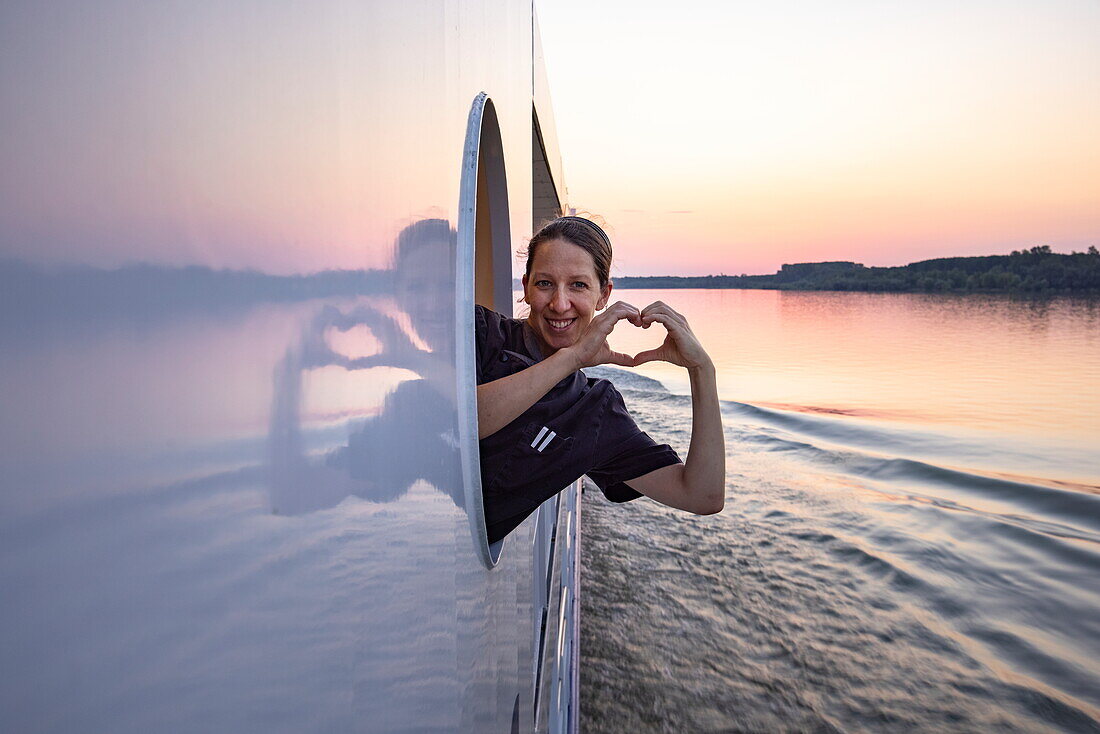 Lächelnde Frau schaut durch Fensteröffnung an Bord von Flusskreuzfahrtschiff Maxima (nicko cruises) auf der Donau und formt mit ihren Händen ein Herz bei Sonnenuntergang, Rumänien, Europa