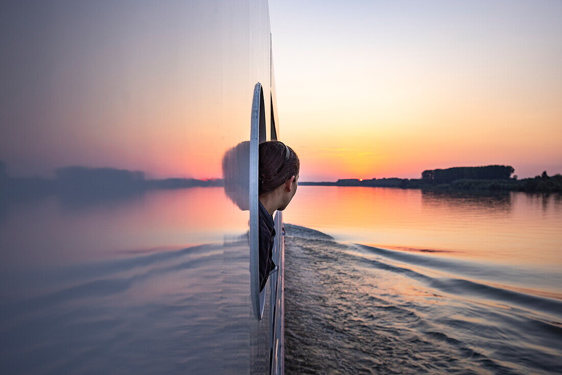 Frau schaut durch Fensteröffnung an Bord von Flusskreuzfahrtschiff Maxima (nicko cruises) auf der Donau bei Sonnenuntergang, in der Nähe von Golubac, Rumänien, Europa