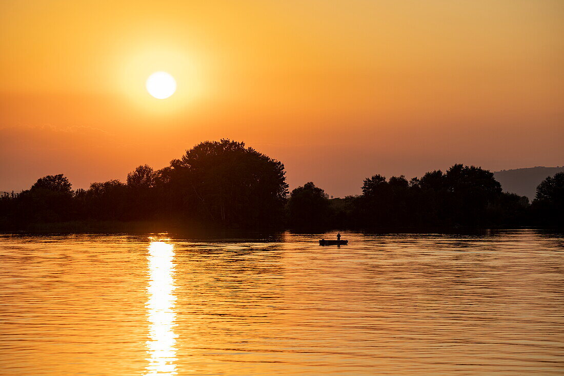 Silhouette von Fischer in Schlauchboot auf der Donau bei Sonnenuntergang, in der Nähe von Coronini, Caraș-Severin, Rumänien, Europa