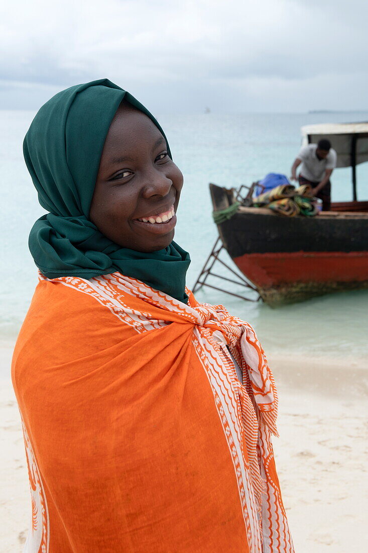  Smiling local woman in orange clothing with boat on the beach, near Stonetown, Zanzibar City, Zanzibar, Tanzania, Africa 