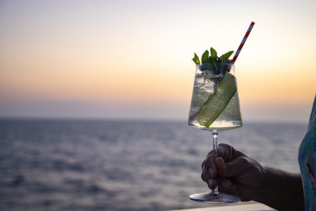 Hand hält Gin-Tonic-Cocktail an der Reling des Expeditionskreuzfahrtschiffes SH Diana (Swan Hellenic) im Roten Meer bei Sonnenuntergang, auf See, in der Nähe von Saudi-Arabien, Arabische Halbinsel