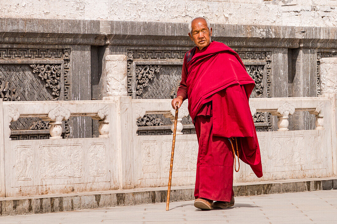  Tibetan monk in red robe with stick walks along an ornate wall of Kumbum Champa Ling Monastery, Xining, China 