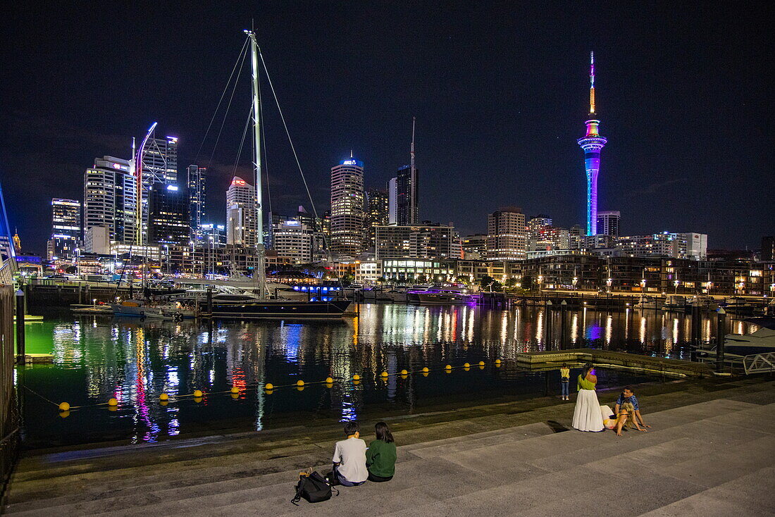  People relaxing on steps at Viaduct Basin with skyline and illuminated Sky Tower at night, Auckland, North Island, New Zealand 