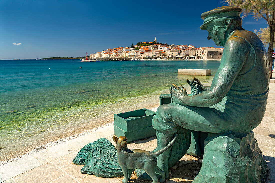  Sculpture of a fisherman on the beach and the peninsula with the old town of Primosten, Croatia, Europe  