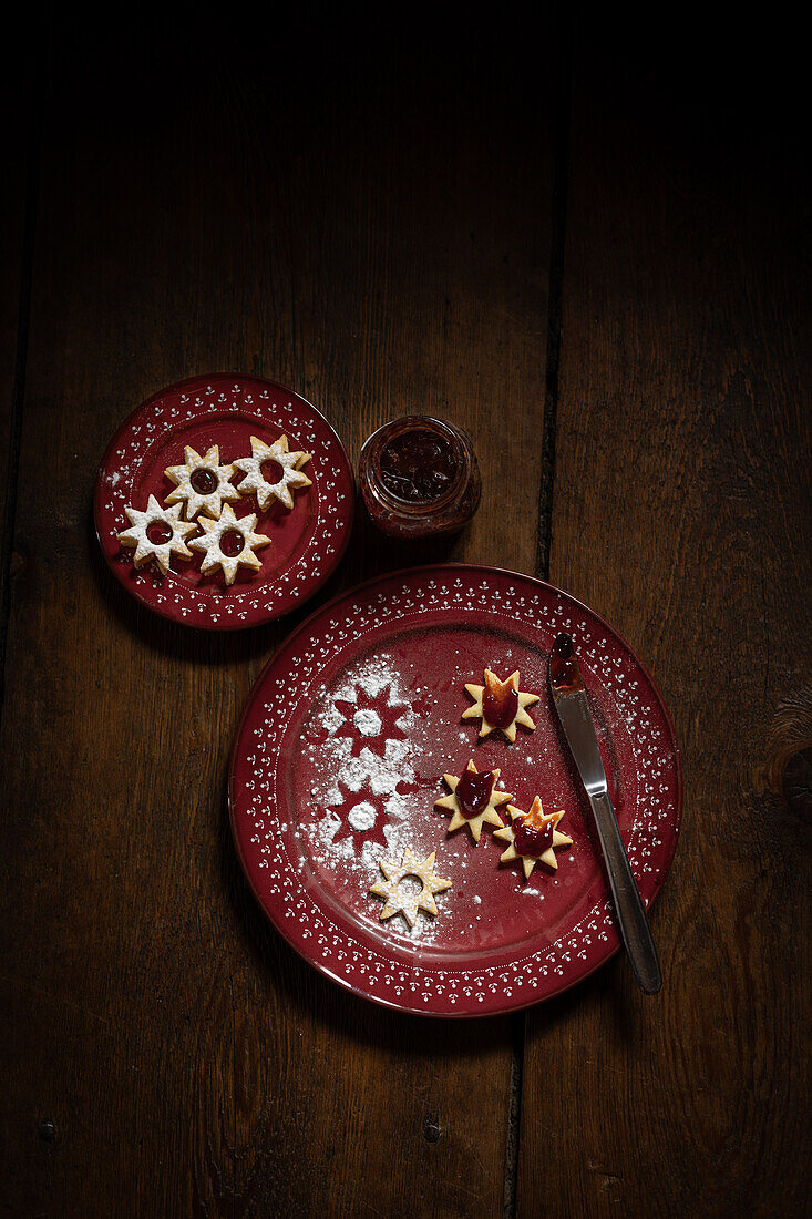 Weihnachtsplätzchen (Spitzbuben) mit Marmelade und Messer, rote Teller, auf Holzuntergrund