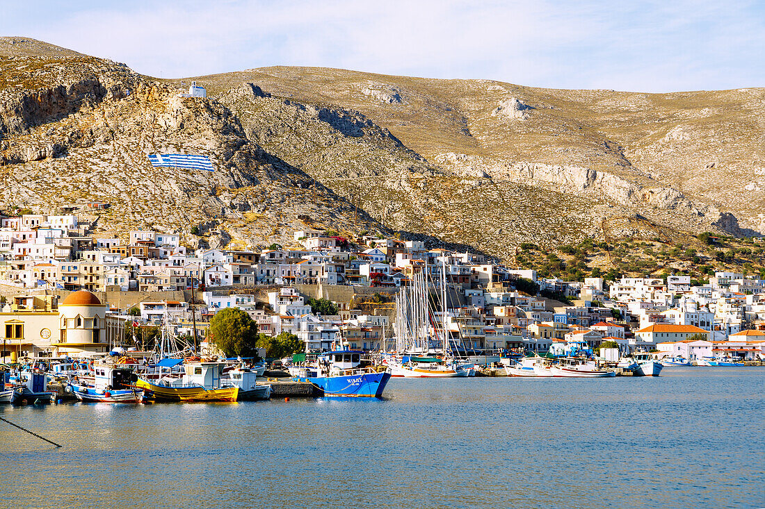  Island capital Póthia, white chapel, Greek flag, pastel-colored houses and boats in the harbor on the island of Kalymnos (Kalimnos) in Greece 