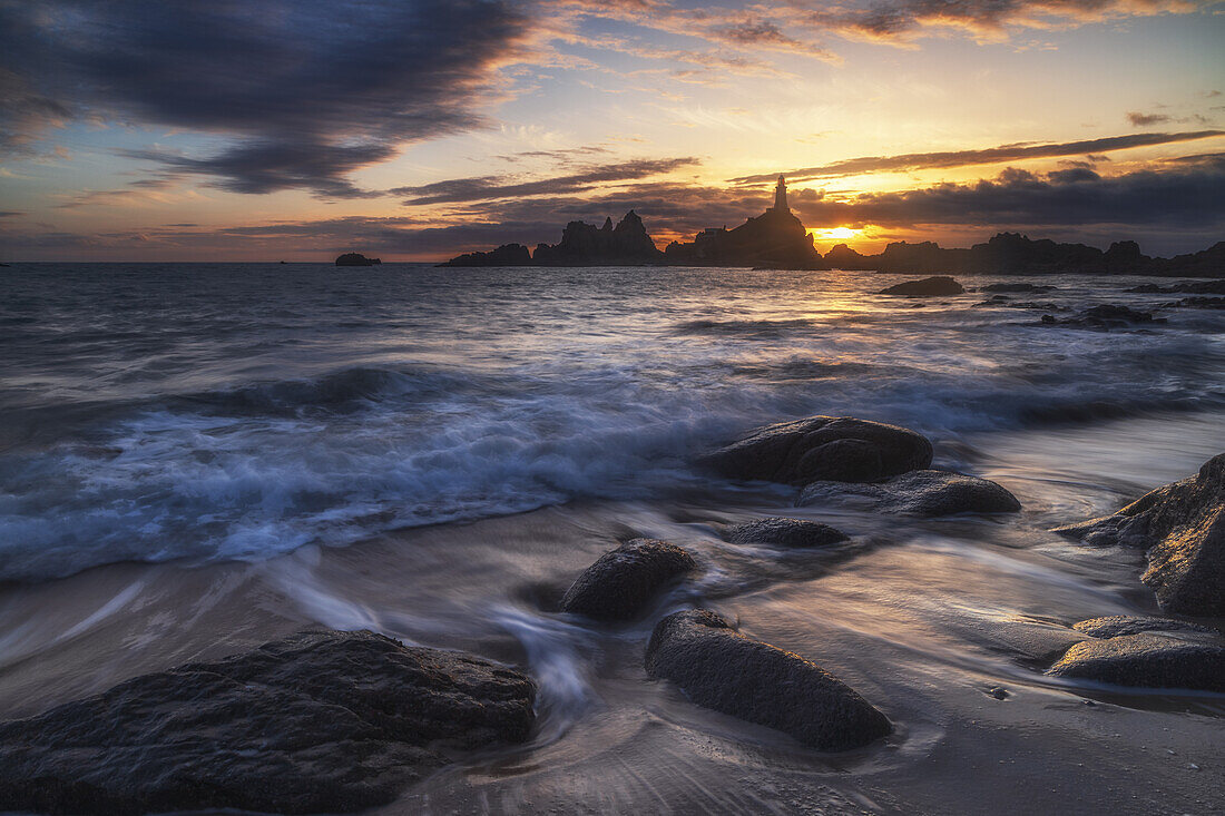  Le Corbiere lighthouse at sunset during low tide. Waves and stones on the beach. 