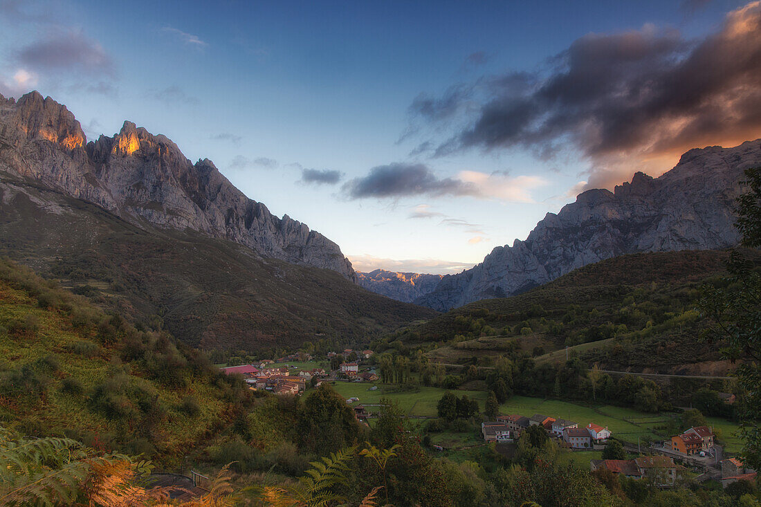  Morning view of the mountain village of Posada de Valdeon. Mountain peaks illuminated by the sun. Ferns in the foreground. 