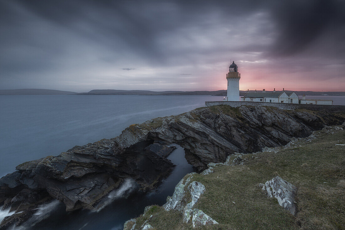  Kirkabister lighthouse stands on a rocky arch by the sea. Red sunset, blue sky. 