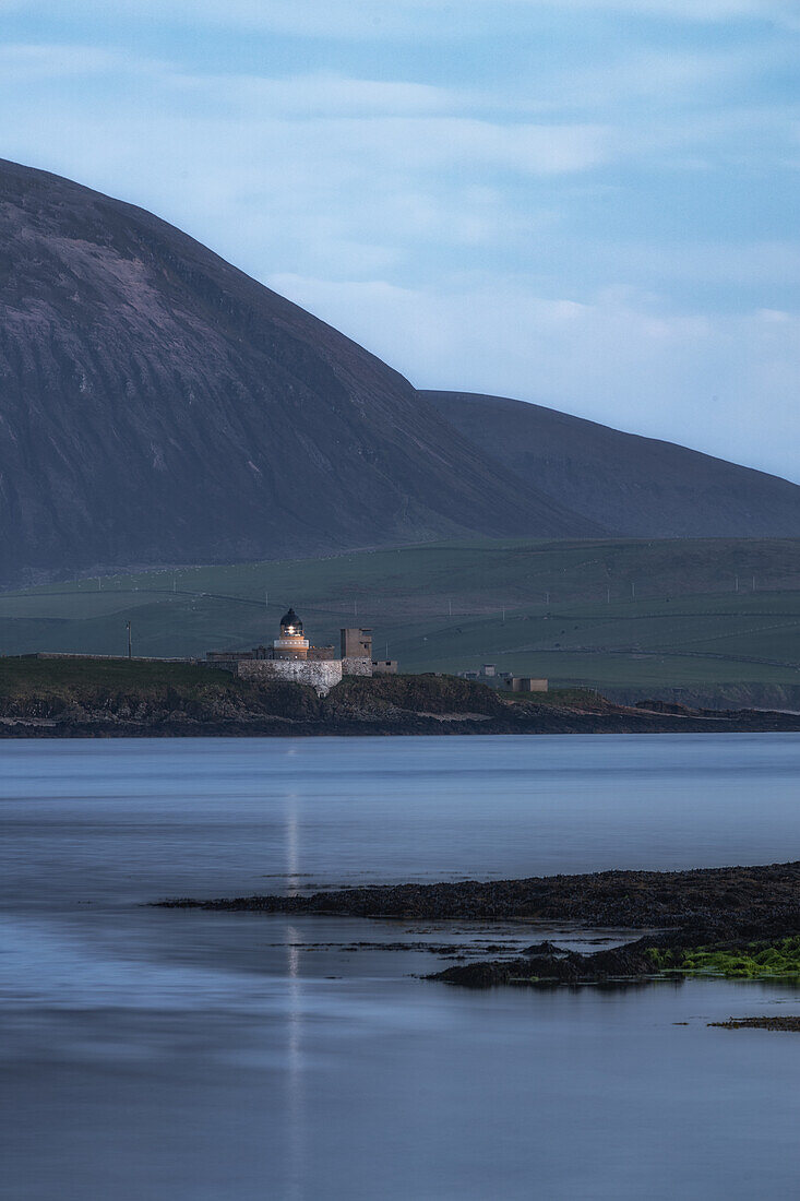  Graemsey low lighthouse in the evening light in front of mountains with lights on. Calm sea in the foreground. 