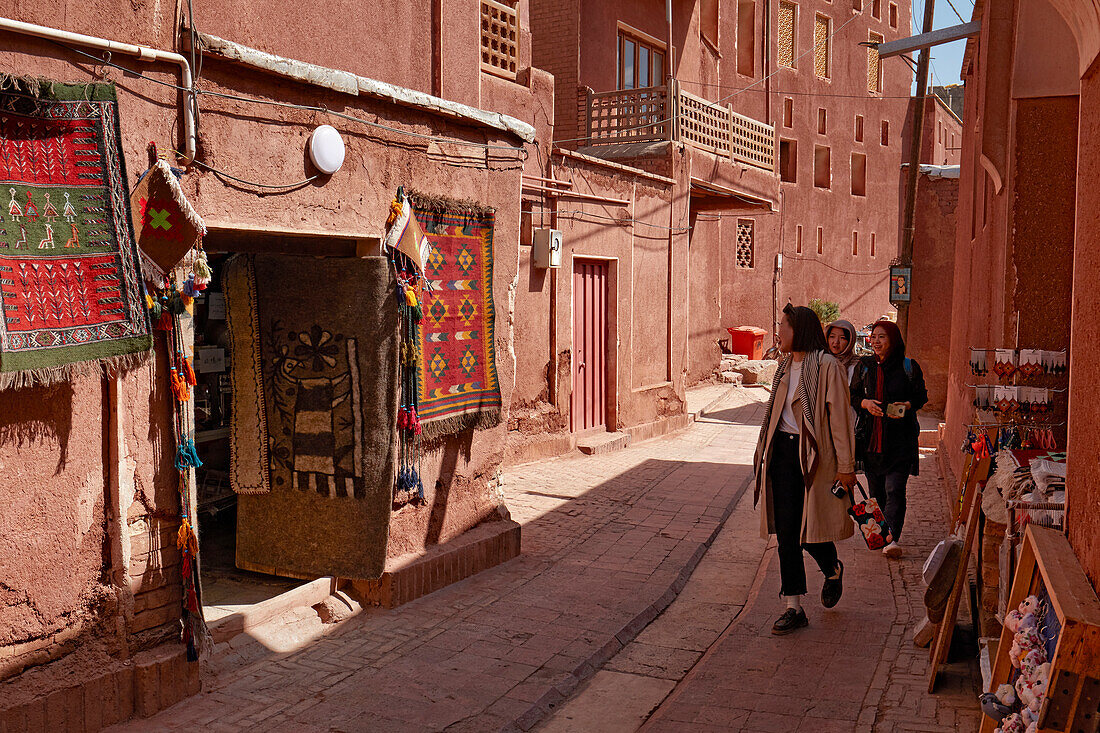  Touristen kommen an einem Souvenirladen vorbei, vor dem farbenfrohe traditionell gewebte Teppiche ausgestellt sind. Dorf Abyaneh, Kreis Natanz, Iran. 