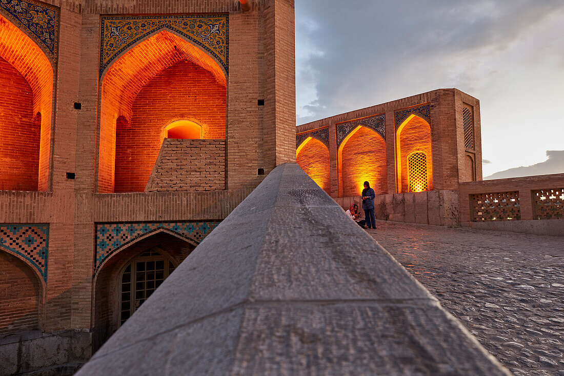 View of illuminated vaulted arches of the 17th century Khaju Bridge on Zayanderud river at dusk. Isfahan, Iran.