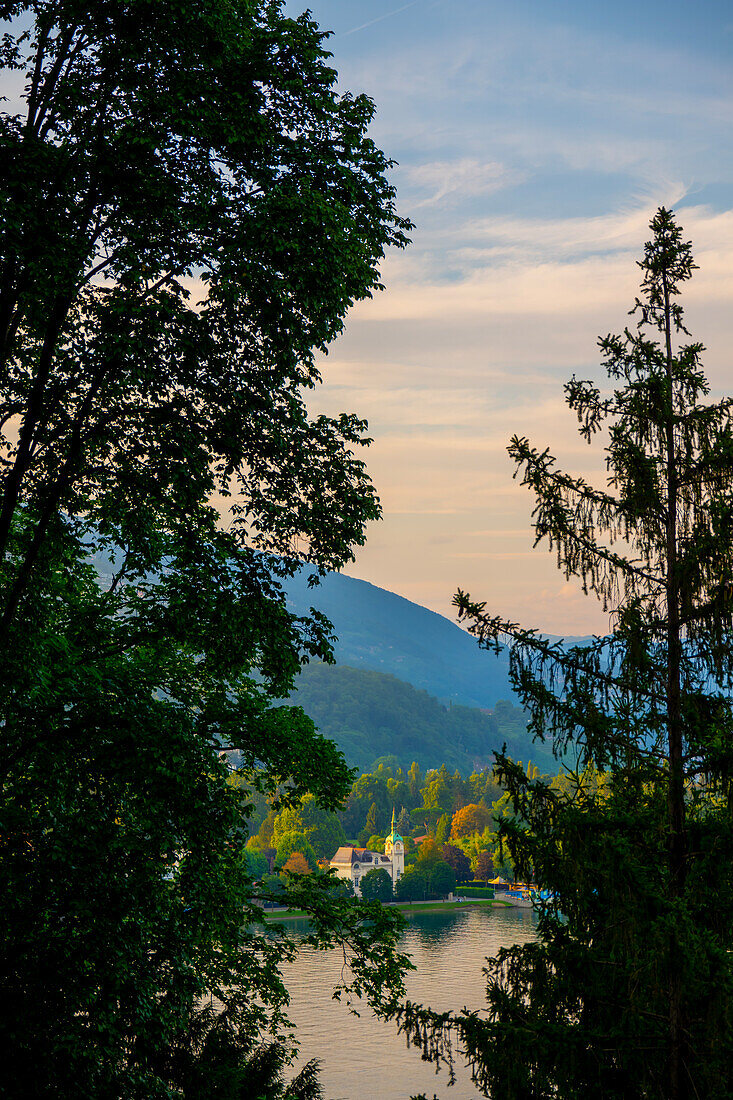  Panoramablick über ein Haus mit Turm am Luganersee mit Bergen und blauem Himmel und Wolken im Sonnenuntergang in Caslano, Tessin, Schweiz. 