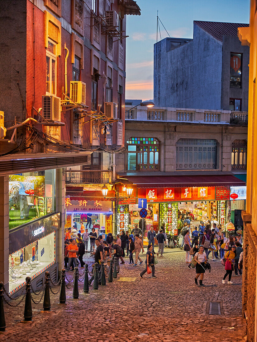 People walk in Rua de S. Paulo (Dasanba) street illuminated at dusk. Macau, China.