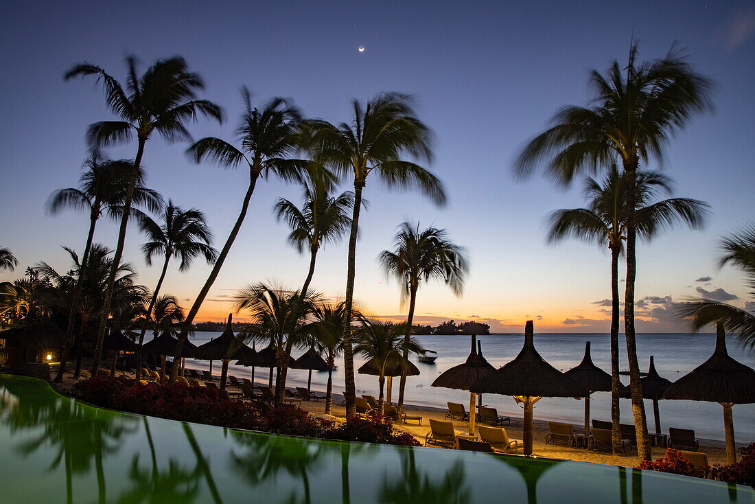 Strohschirme mit Kokospalmen am Strand bei Sonnenuntergang, Royal Palms Beachcomber Resorts, Grand Baie, Rivière du Rempart, Insel Mauritius, Indischer Ozean