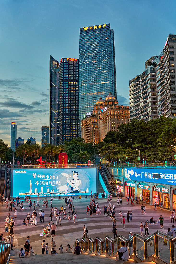  Menschen gehen auf dem Huacheng-Platz spazieren, der von modernen, in der Abenddämmerung beleuchteten Hochhäusern umgeben ist. Guangzhou, Provinz Guangdong, China. 