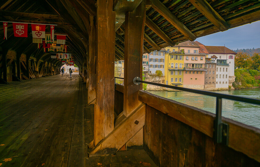  Innen- und Außenseite der alten historischen überdachten Holzbrücke mit Flaggen und Haus über der Aare an einem sonnigen Tag in Olten, Kanton Solothurn, Schweiz. 