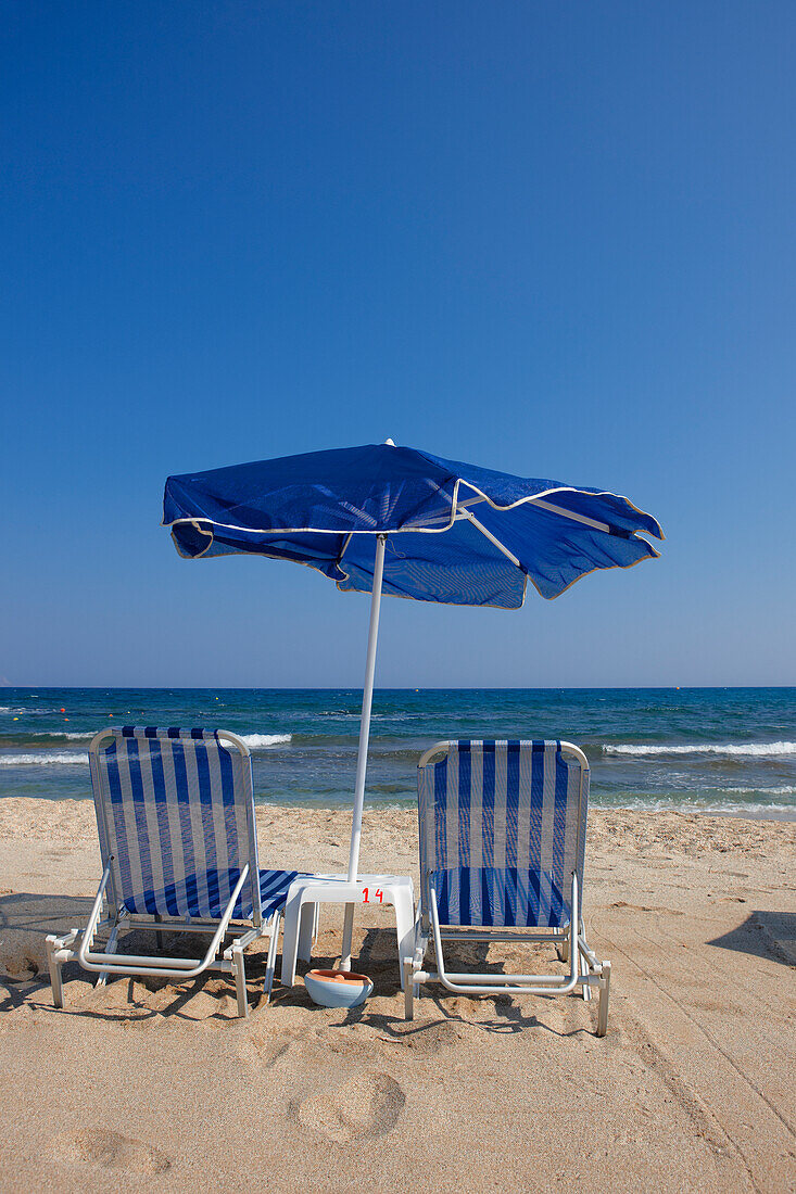  Zwei Liegestühle unter einem blau-weißen Sonnenschirm an einem Sandstrand in der Nähe von Hersonissos, Kreta, Griechenland. 