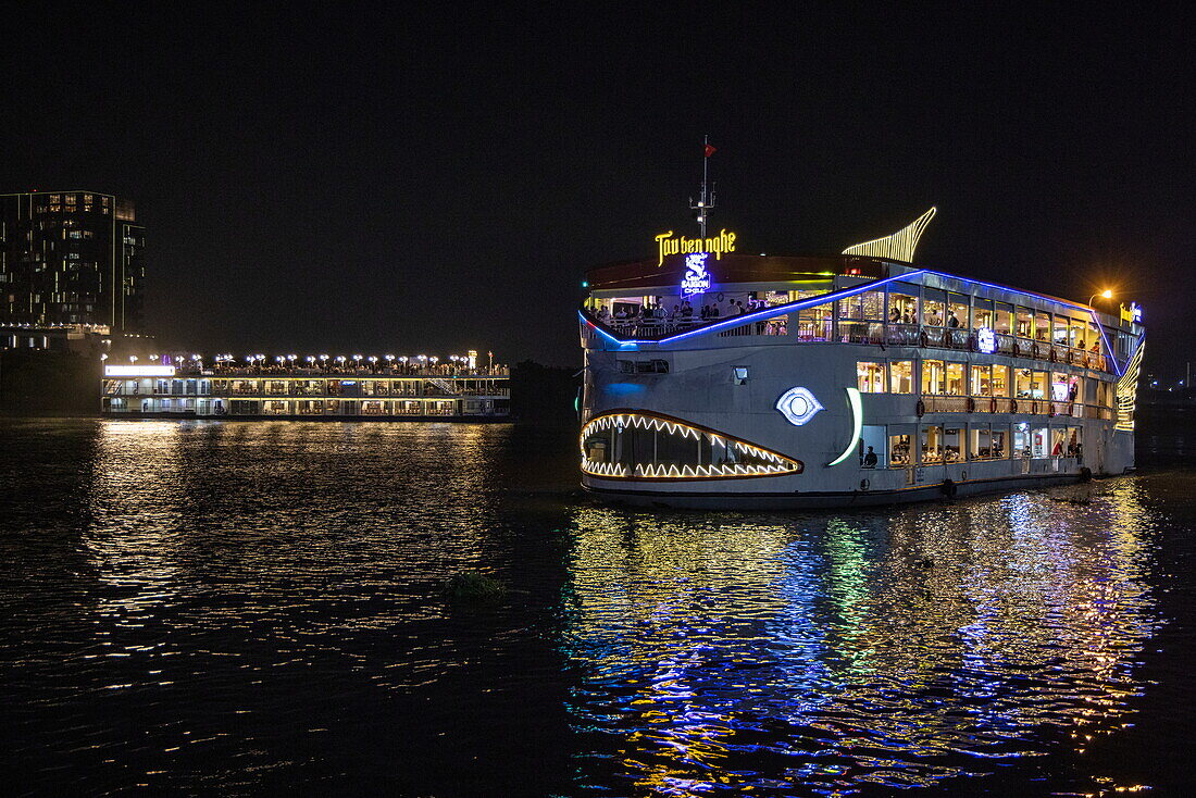  Illuminated dinner cruise boat with shark mouth lights approaches pier on Saigon River at night, Ho Chi Minh City, Vietnam, Asia 