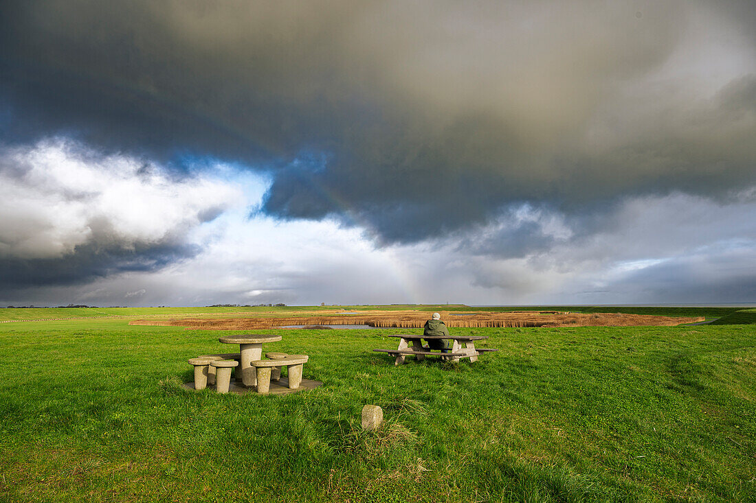 A person sitting at a picnic table in a vast green field under a dramatic cloudy sky.