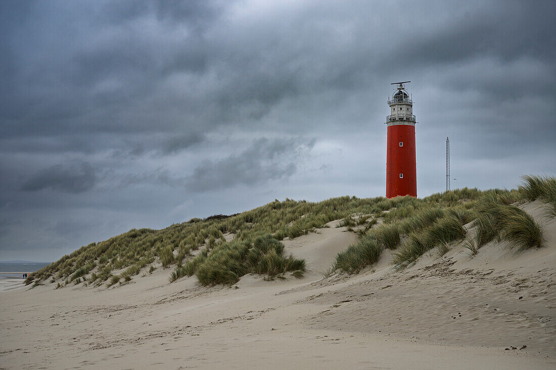 A red lighthouse stands on a sandy dune under a cloudy sky.