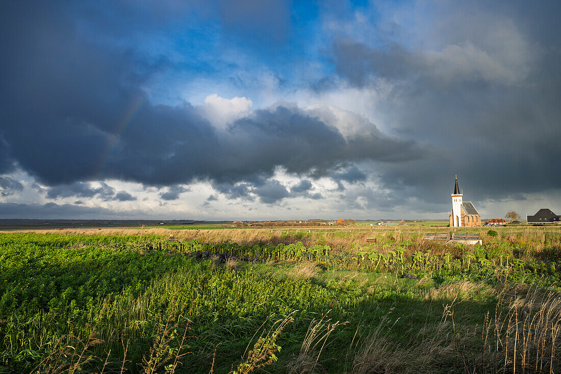 A rural landscape with a church under a dramatic cloudy sky, surrounded by green fields.