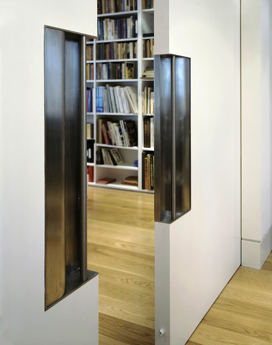 A half open sliding door with an integrated, stainless steel handle and a view of a bookshelf