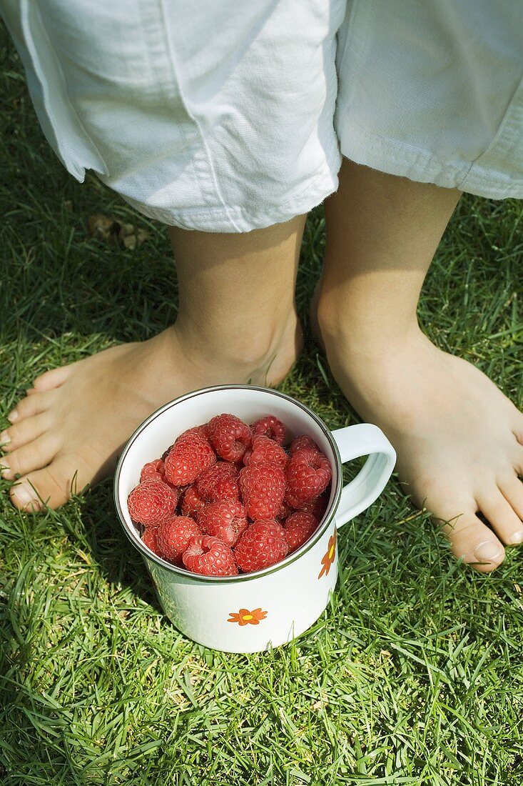 Raspberries in a mug on grass between a child's feet