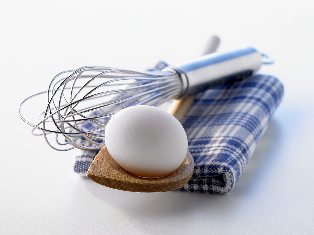 A white egg on a wooden spoon and an egg whisk